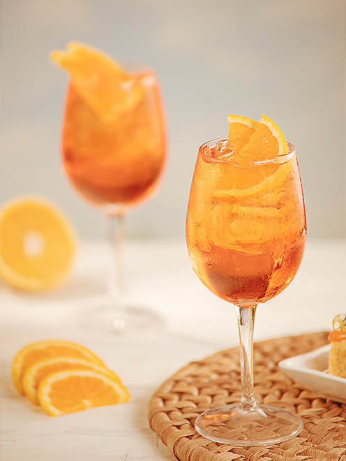 Close-up em duas taças de Aperol Spritz com gelo e fatias frescas de laranja, sobre um descanso de mesa artesanal em palha trançada.