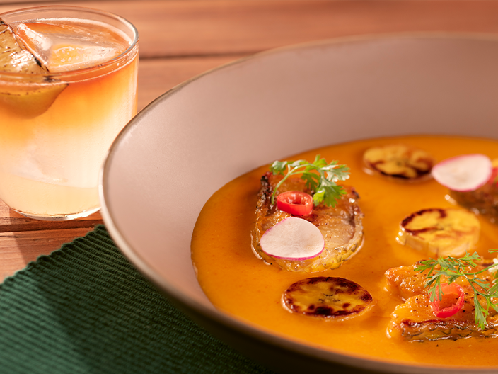 Prato de peixe grelhado servido em molho cremoso alaranjado e ervas frescas, acompanhado de bebida cítrica servida em copo de vidro sobre mesa de madeira.
